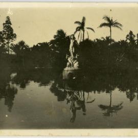Photo of a statue above a still pond flanked by palms. Location unknown