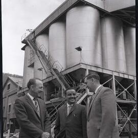 Three men in suits outside a refinery, 1963