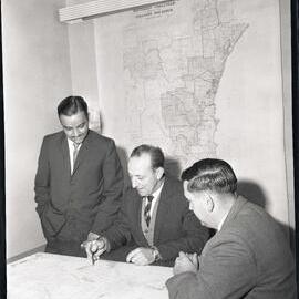 Three men at a desk reading a plans, 1963