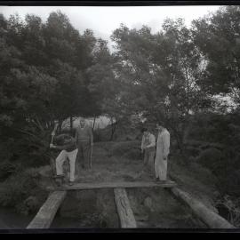 Four men standing on wooden logs in bushland, 1963