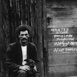 William Kelly sitting outside a hut. Sign: "WANTED a cook. Young Lady preferred. Apply at once. If not sooner."