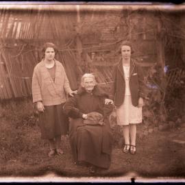 Older woman in chair with two young women standing, c. 1910s