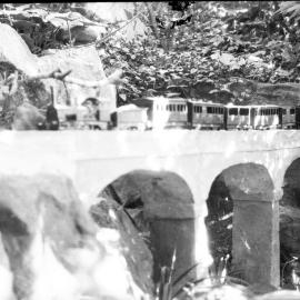 Model locomotive and passenger carriages, on model viaduct in garden setting, unknown date and location.