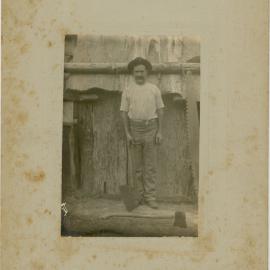 A portrait of a gentleman, a timber worker. He is wearing a hat and holding an axe. In front of him a axe is displayed and on the right side a big tree saw, [n.d.]