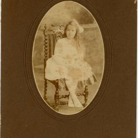 Teenaged girl sitting on a carved chair. She wears a light coloured dress and has a big bow in her long dark coloured hair, [n.d.]