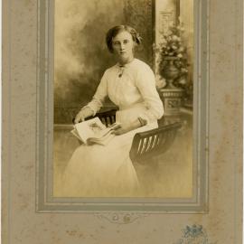 Portrait of a young lady holding a magazine in her hands. She is sitting on a half round shaped stool and  wearing a light coloured dress, [n.d.]